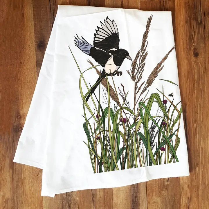White fabric with a print of a magpie flying among tall grasses and reeds on a wooden floor surface.