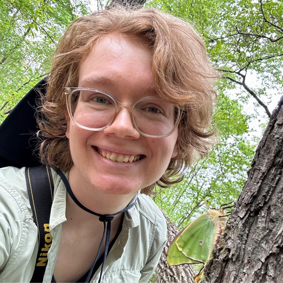 Smiling woman outdoors with glasses and a backpack strap, a green butterfly rests on a tree trunk beside her.